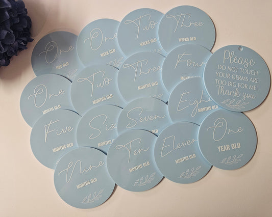 Round blue baby milestone discs with white script text showing weekly, monthly, and one-year age markers, plus “Please do not touch” disc.