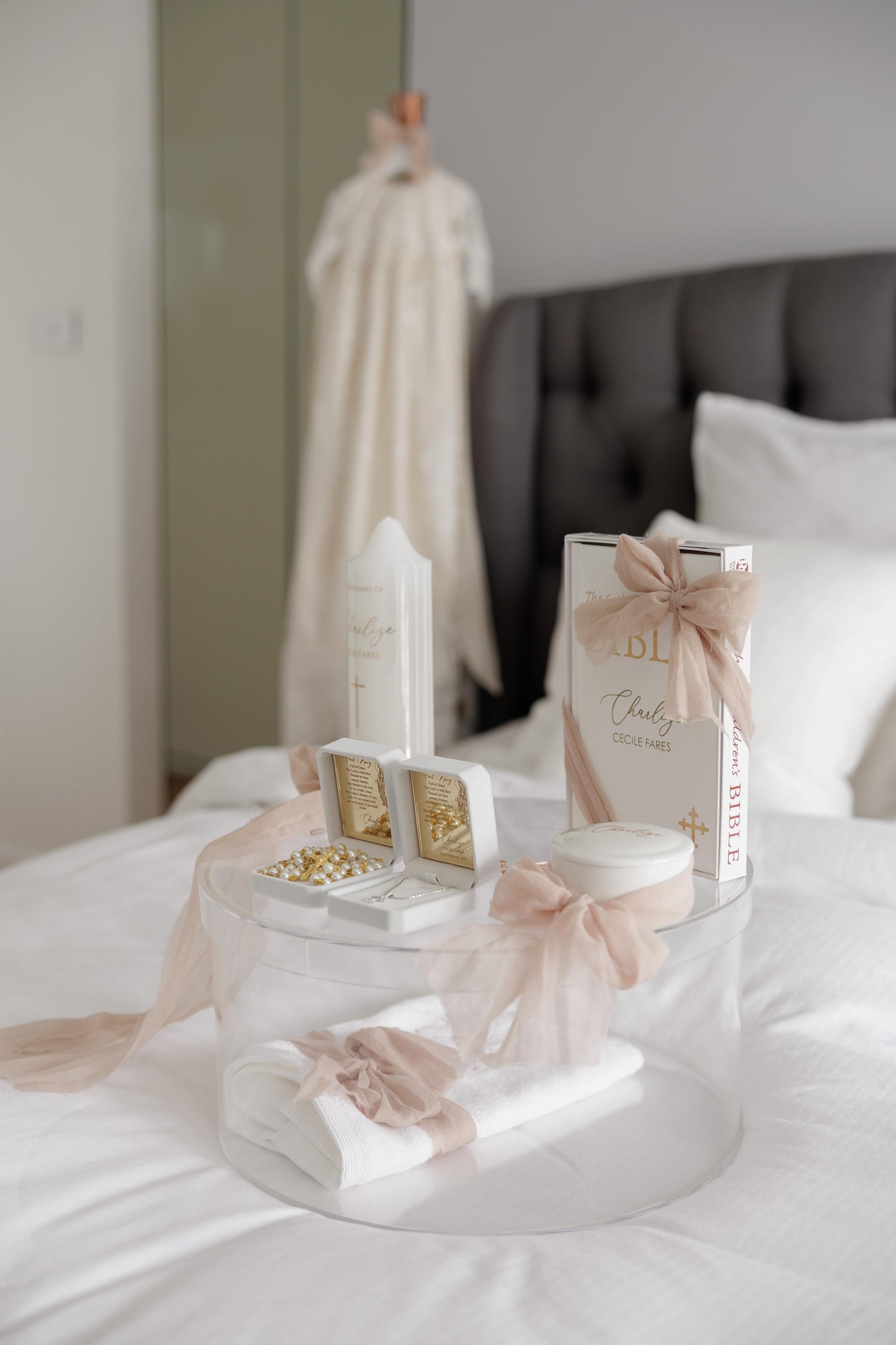 A beautifully arranged Christening gift set with a personalized candle, decorative Bible, jewelry, keepsake box, and towel in a clear christening box, with a gown in the background.