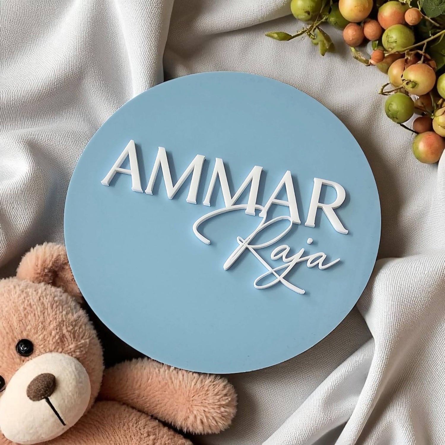 Round personalised newborn name plaque with white script, blue background, teddy bear and berry accents – custom colours and fonts available.