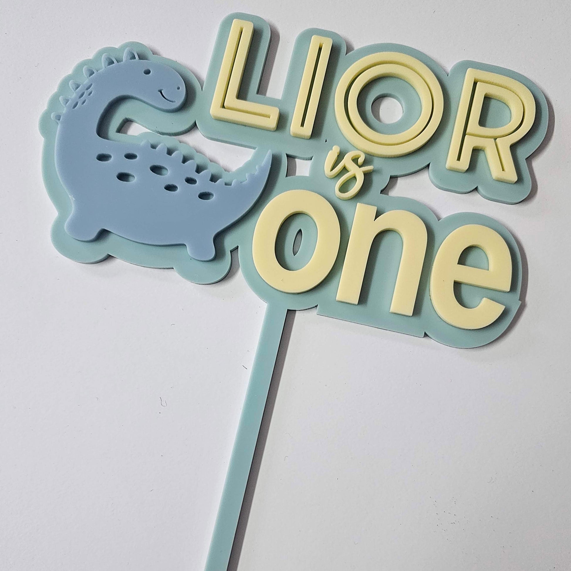 Dinosaur-themed cake topper with "Lior is One" in pastel blue and yellow, featuring a smiling blue dino. Perfect for a first birthday celebration.