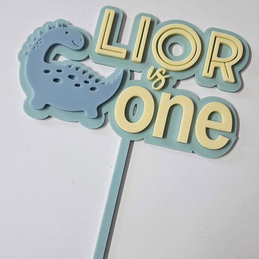 Dinosaur-themed cake topper with "Lior is One" in pastel blue and yellow, featuring a smiling blue dino. Perfect for a first birthday celebration.