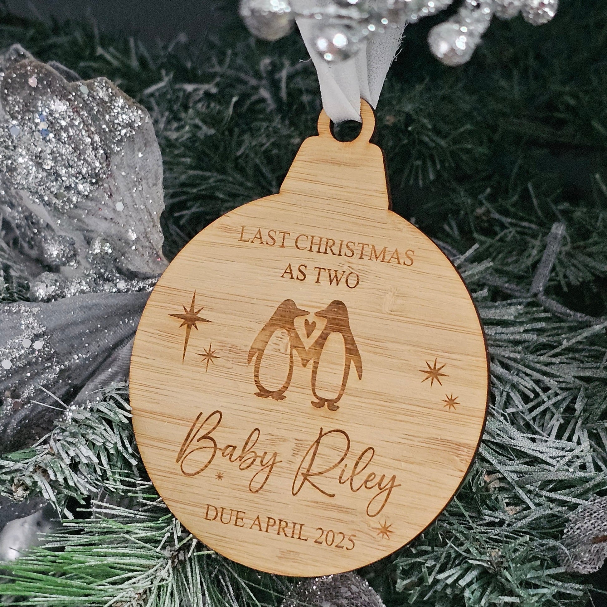 Wooden Christmas ornament engraved with two penguins, heart, stars, and personalised baby announcement text, hanging on a decorated tree.