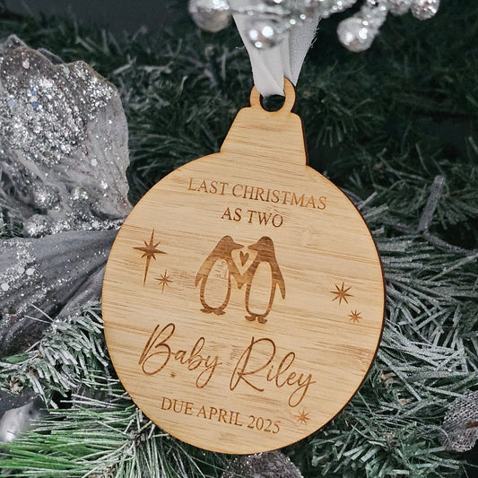 Wooden Christmas ornament engraved with two penguins, heart, stars, and personalised baby announcement text, hanging on a decorated tree.