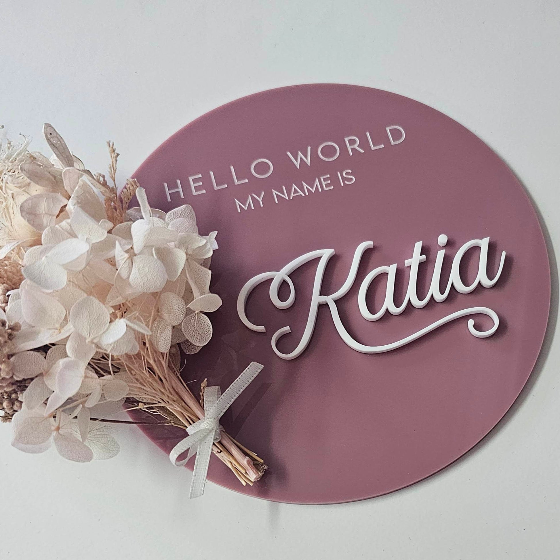Acrylic baby name sign in mauve with 'Hello World My Name Is' engraved text in white and raised Acrylic cursive name, featuring dried flower bundle tied with white ribbon