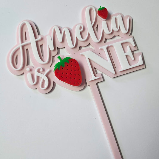 Pink and white cake topper with personalised name  decorated with cute red strawberries. Perfect for a first birthday celebration.