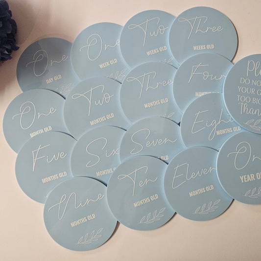 Set of round light blue acrylic baby milestone discs with white text, including age markers from one day to one year and a germ safety message