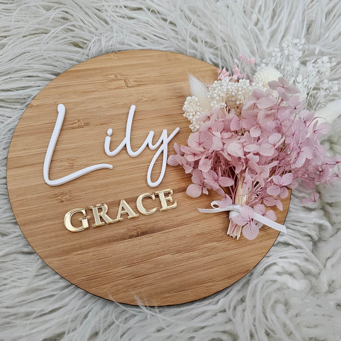 Round bamboo baby name plaque with white and gold acrylic layered lettering and dried flower bunch tied with ribbon.