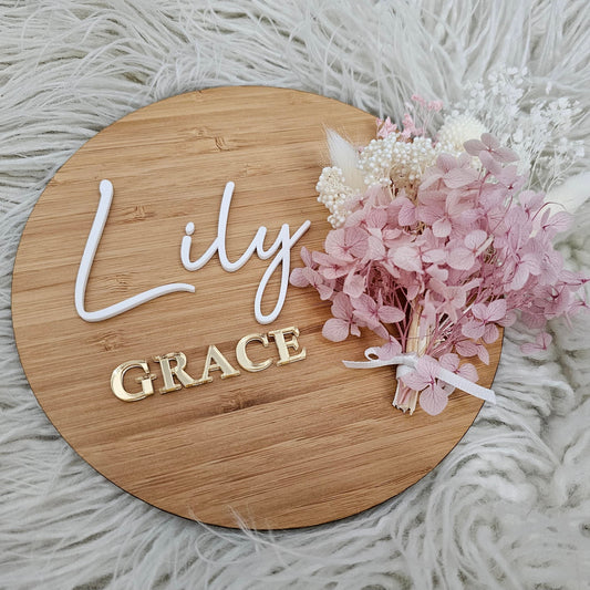 Round bamboo baby name plaque with white and gold acrylic layered lettering and dried flower bunch tied with ribbon.