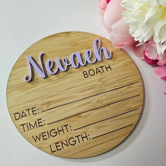 Round wooden baby plaque with 3D script name in pink, engraved surname in white block lettering, and space for birth details.