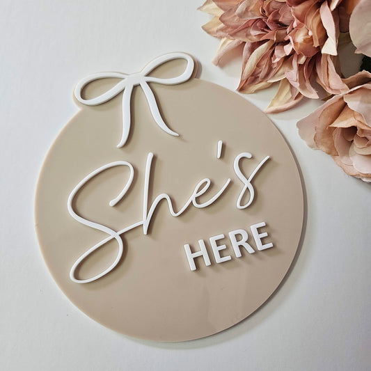 Freestanding round baby plaque with raised 3D “She’s HERE” lettering and soft bow detail in white script and block fonts.