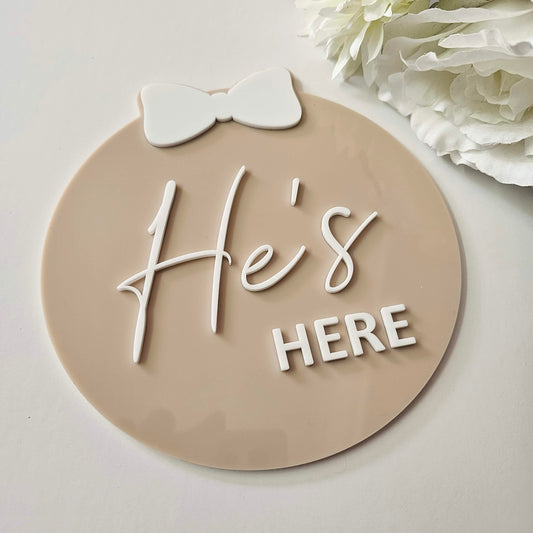 Freestanding round baby plaque with raised 3D “He’s HERE” text in white, featuring a bow accent in script and block font styling.