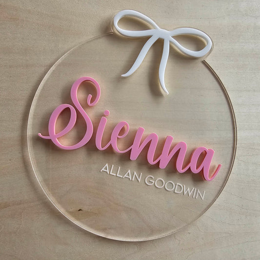 Clear acrylic newborn keepsake plaque with pink and white text, white bow, and layered name design in cursive and block fonts.