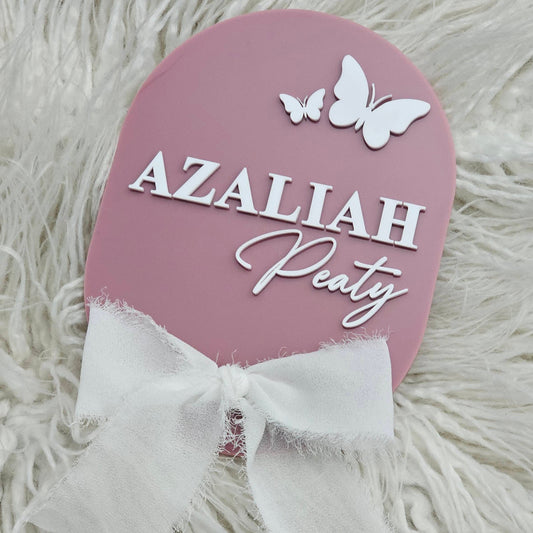 Oval pink personalised newborn keepsake plaque with white lettering, butterfly accents, and a fabric bow – perfect for shelf or surface display.