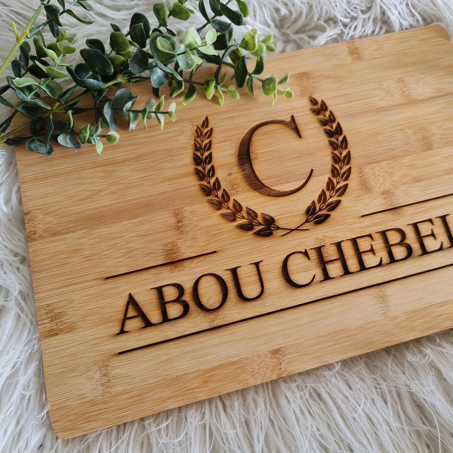 Wooden plaque engraved with monogram “C” inside laurel wreath and custom name “ABOU CHEBEL,” styled on white textured surface with greenery.
