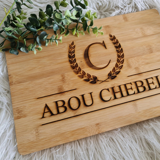Wooden plaque engraved with monogram “C” inside laurel wreath and custom name “ABOU CHEBEL,” styled on white textured surface with greenery.