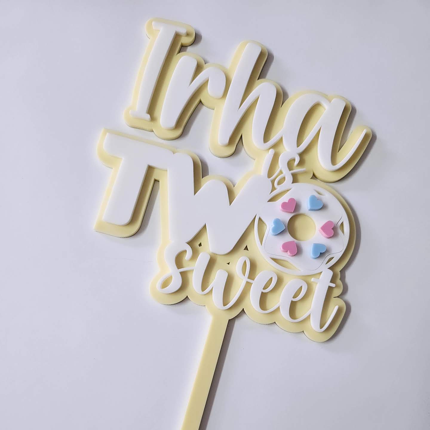 Custom "Irha is Two Sweet" cake topper with pastel yellow and white lettering, featuring a cute donut design with heart sprinkles for a 2nd birthday party.