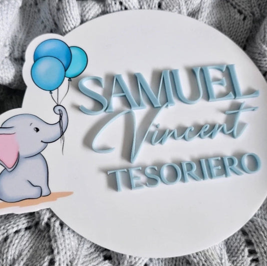 Round baby name plaque with 3D-style blue text and a baby elephant illustration holding two balloons, on soft grey fabric background.