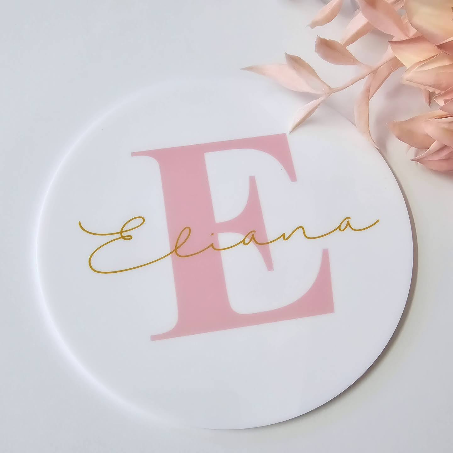 Round baby name plaque with large pink initial, gold script name overlay, and light pink botanical accents – UV printed keepsake plaque.