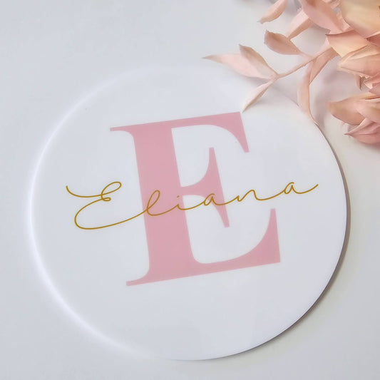 Round baby name plaque with large pink initial, gold script name overlay, and light pink botanical accents – UV printed keepsake plaque.