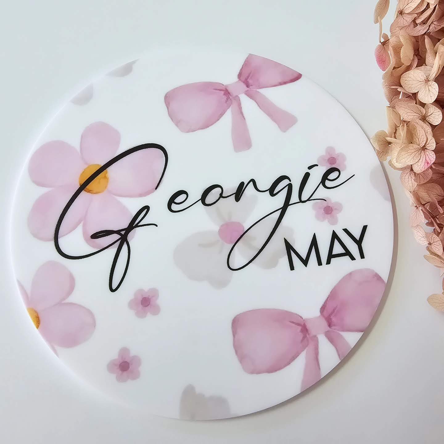 Round baby keepsake plaque with pink personalised text and bow accents in script and block fonts, UV printed for a clean, durable finish.