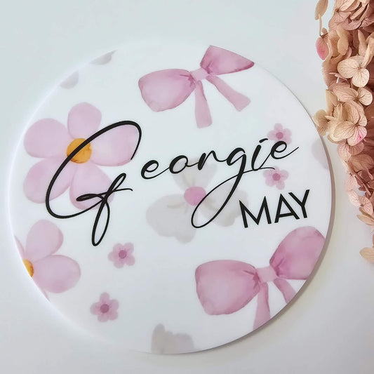 Round baby keepsake plaque with pink personalised text and bow accents in script and block fonts, UV printed for a clean, durable finish.